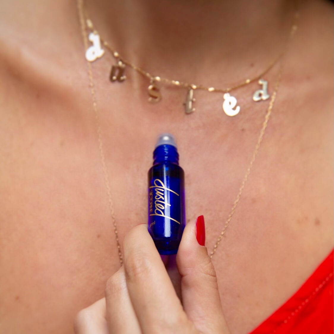 Dusted fragrance in a blue glass bottle with gold letters, held by a hand with red nails, over the décolleté of a white person wearing two gold necklaces, one reads "dusted" in gold charms. 